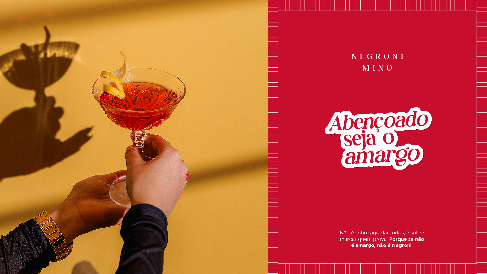 Mão erguendo uma taça de Negroni em parede amarela ao lado do cartaz Abençoado seja o amargo